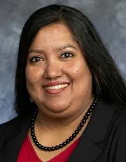 A portrait of a woman with long, straight black hair and medium brown skin. She is smiling warmly and wearing a black blazer over a red top, accessorized with a black beaded necklace. The background is a textured gray-blue, giving the image a formal and professional appearance. Her expression is friendly and approachable.