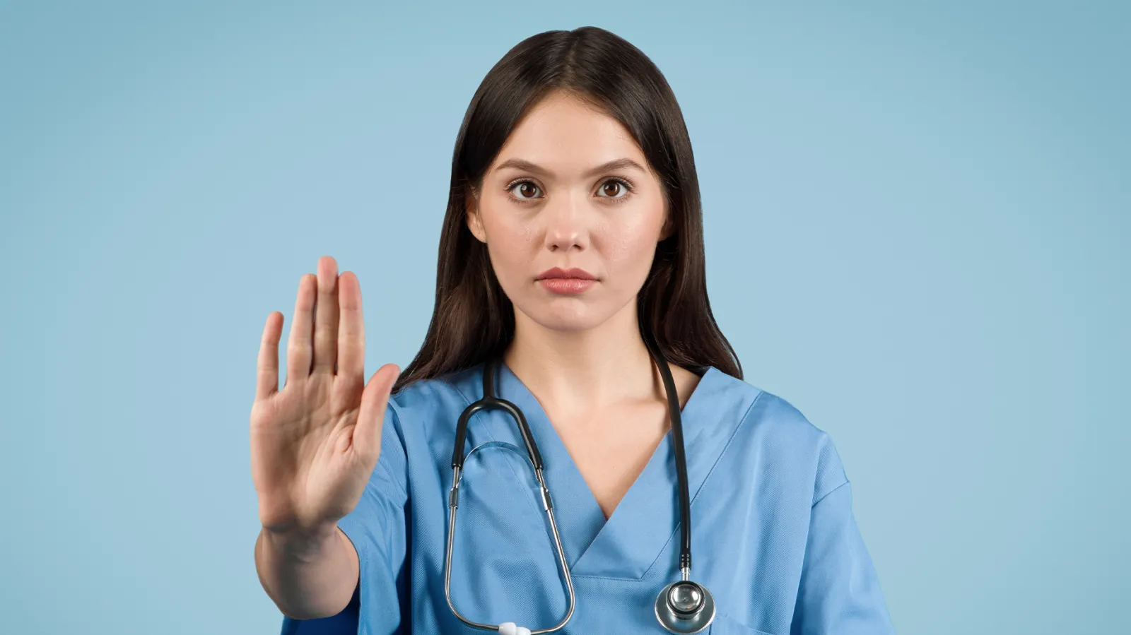 Female healthcare professional in blue scrubs with a stethoscope around her neck, raising her hand in a 'stop' gesture against a light blue background