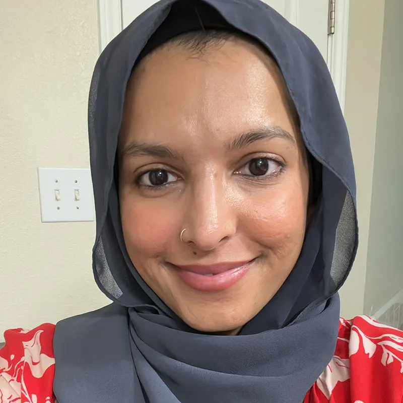 A portrait of a woman with medium brown skin wearing a gray hijab, which frames her face. She has a gentle smile and a small nose ring on her left nostril. Her eyes are warm brown, and her eyebrows are well-groomed. She is wearing a red blouse with a white floral pattern. The background shows an indoor setting with a light switch and a white door, giving a casual, home-like atmosphere. Her expression is friendly and approachable.