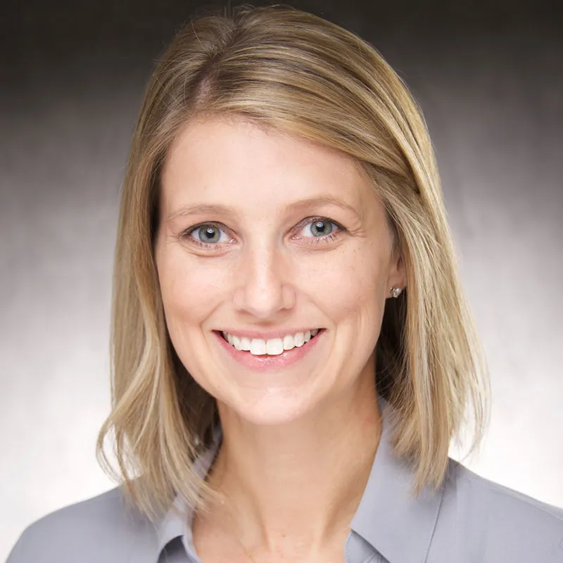 A portrait of a woman with shoulder-length blonde hair, blue eyes, and light skin. She is smiling broadly, showing her teeth, and wearing a light gray blouse with a collar. The background features a gradient from dark gray to light gray, creating a soft, neutral backdrop. Her expression is friendly and professional.