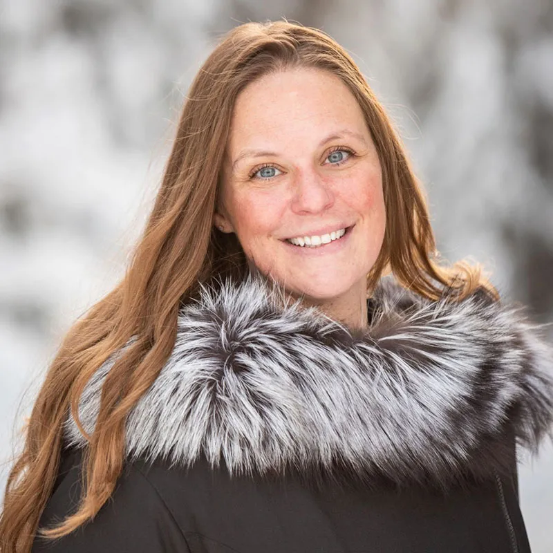 A portrait of a smiling woman with long, straight, light brown hair and blue eyes. She is outdoors with a snowy, blurred background. She is wearing a black winter coat with a large, fluffy faux fur collar in gray and white tones. Her expression is cheerful, and her rosy cheeks suggest a cold, winter setting.