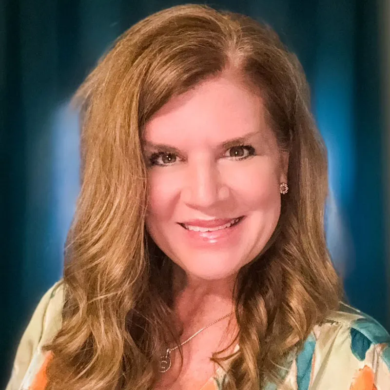 A smiling woman with long, wavy light brown hair poses in front of a softly lit teal background. She is wearing a colorful floral top, a silver necklace, and stud earrings.