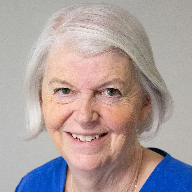 A portrait of an older woman with fair skin and short, neatly styled white hair. She is wearing a bright blue blouse and smiling warmly, showing her teeth. Her green eyes are slightly crinkled at the edges. The background is a plain, light gray color. Her expression conveys kindness and approachability.