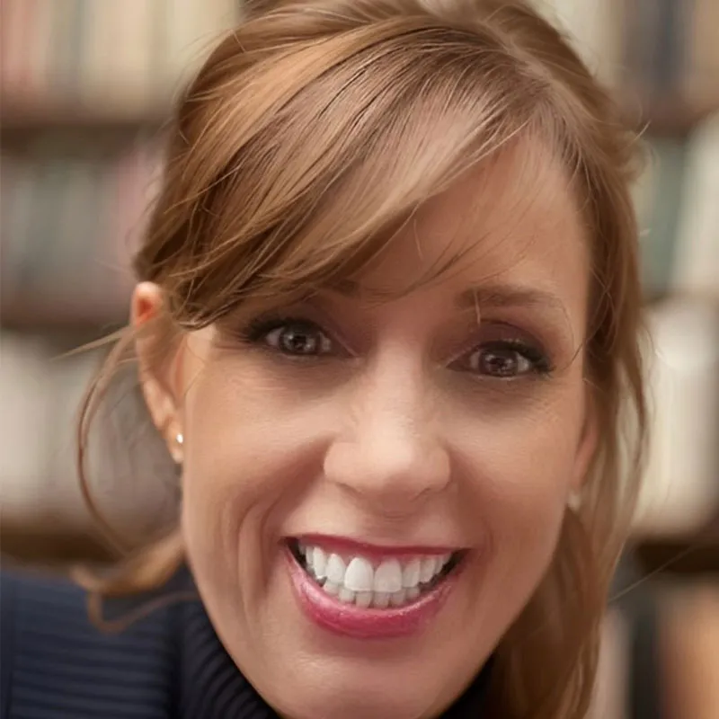 A close-up portrait of a smiling woman with light brown hair styled with bangs swept to the side and subtle waves. She has fair skin, bright eyes, and a wide smile showing her teeth. She is wearing a dark blue turtleneck sweater. The background features slightly blurred bookshelves, suggesting a library or study setting. Her expression is cheerful and friendly.