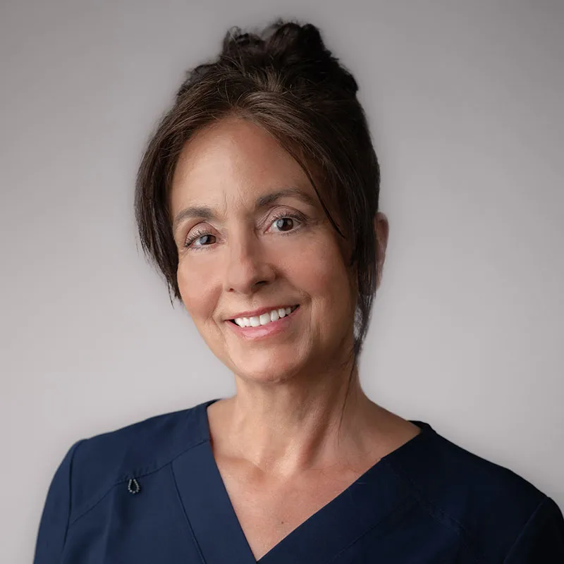 A portrait of a woman with medium-length brown hair styled in a loose updo. She has a warm smile, brown eyes, and light skin. She is wearing a dark navy-blue top, possibly a scrub or uniform. The background is a soft, neutral gray. Her expression is friendly and approachable, conveying a professional and caring demeanor.