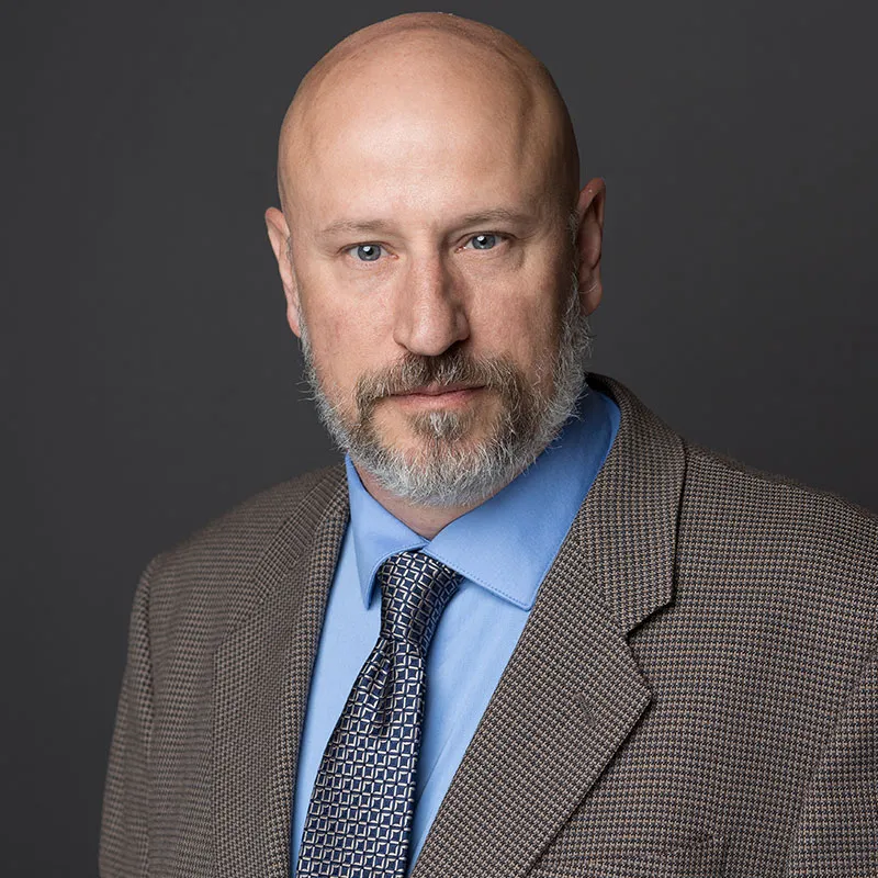 A portrait of a bald man with a neatly trimmed gray beard and blue eyes. He is wearing a brown houndstooth-patterned blazer, a light blue dress shirt, and a patterned blue tie. His expression is serious and confident, with a direct gaze at the camera. The background is a solid dark gray, providing a professional and formal setting.