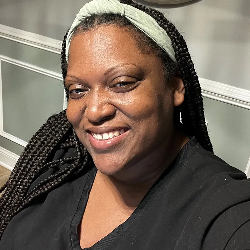 A portrait of a smiling woman with dark brown skin and long braided hair. She is wearing a black top and a light green headband. Her expression is warm and friendly, with her eyes slightly squinting from her smile. The background consists of an indoor setting with gray walls featuring white trim. The lighting is soft and natural, giving the photo a relaxed and approachable feel.