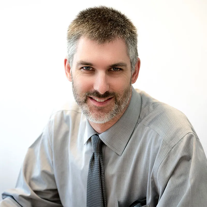 A portrait of a middle-aged man with short, salt-and-pepper hair and a neatly trimmed beard. He is wearing a light gray dress shirt and a gray tie with subtle polka dots. His expression is warm and confident with a slight smile. The background is a plain white surface, creating a clean and professional setting.