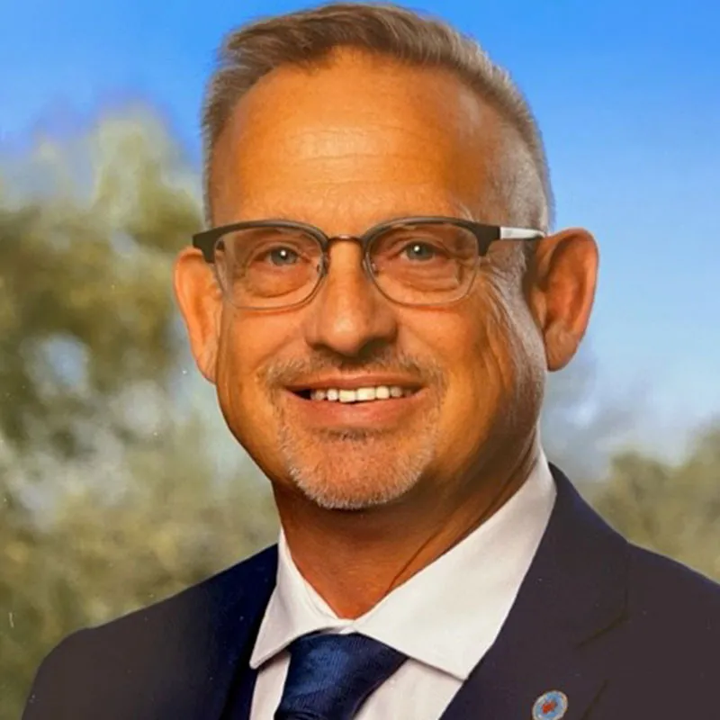 A portrait of a middle-aged man with short, light brown hair that is graying at the sides, neatly styled. He has a trimmed goatee and wears rectangular glasses with a thin, dark frame. He is dressed in a navy blue suit, white dress shirt, and a dark blue tie. The background shows a slightly blurred outdoor setting with green trees and a blue sky. He is smiling warmly, conveying confidence and friendliness.