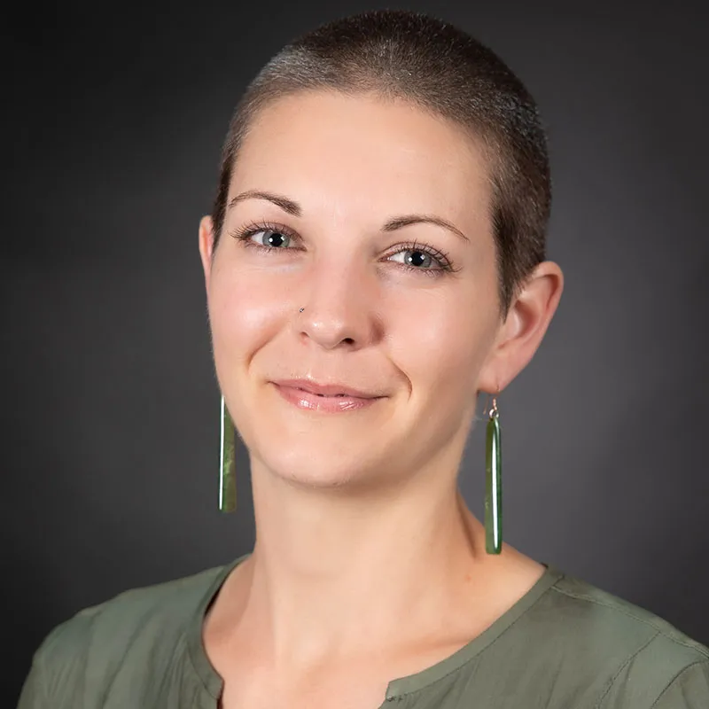 A portrait of a woman with a shaved head, light skin, and a gentle smile. She is wearing long green dangling earrings and an olive-green blouse. Her expression is calm and confident, with her eyes looking directly at the camera. The background is a solid dark gray, providing a professional and neutral setting. Her makeup is minimal, highlighting her natural features.