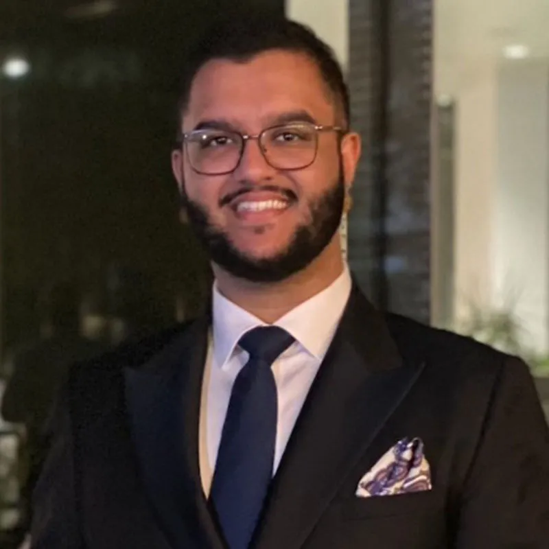 A portrait of a man with short black hair, a neatly trimmed beard, and glasses with a thin black frame. He is wearing a black tuxedo with a white dress shirt, a dark blue tie, and a matching pocket square with a subtle pattern. His expression is cheerful, with a broad smile. The background features a dimly lit modern interior with large glass windows or doors, suggesting an upscale setting.