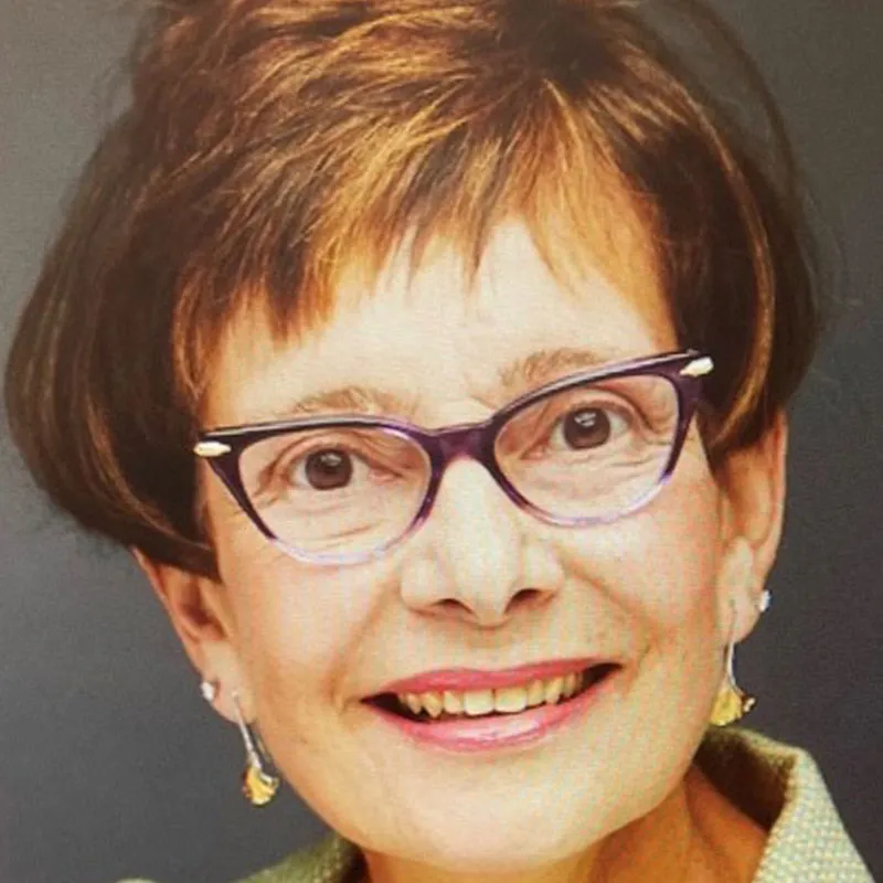 A portrait of a middle-aged woman with short, dark brown hair highlighted with lighter brown streaks. She is wearing purple-framed glasses and dangling gold earrings. Her expression is cheerful with a bright smile showing her teeth. She has fair skin, and the background is a solid dark gray. She is dressed in a muted green blazer or blouse, adding a professional touch to her appearance.