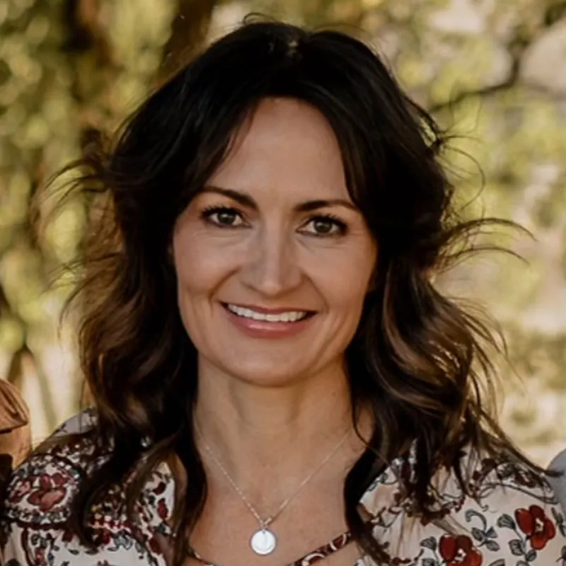 A portrait of a woman with long, wavy dark brown hair and light skin. She is smiling warmly, showing her teeth. She is wearing a floral-patterned blouse with red and beige tones and a silver necklace with a round pendant. The background consists of blurred greenery and trees, suggesting an outdoor setting. Her expression is friendly and confident.