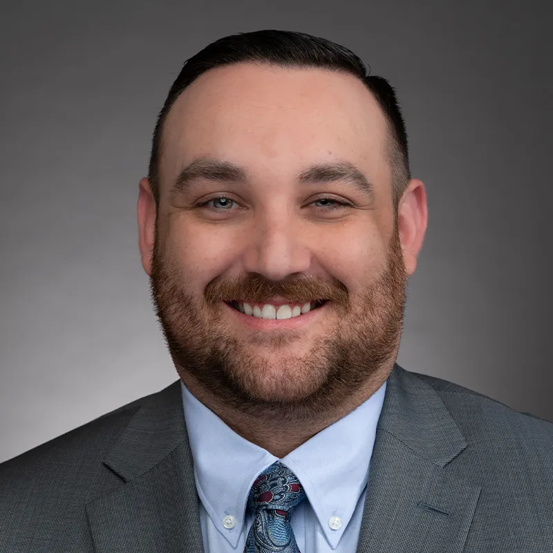 A portrait of a man with short, dark brown hair, a trimmed beard, and light skin. He is smiling warmly and wearing a gray suit jacket, a light blue dress shirt, and a patterned blue tie with paisley and red accents. The background is a neutral gray gradient, giving the image a professional and polished appearance. His expression is approachable and confident.