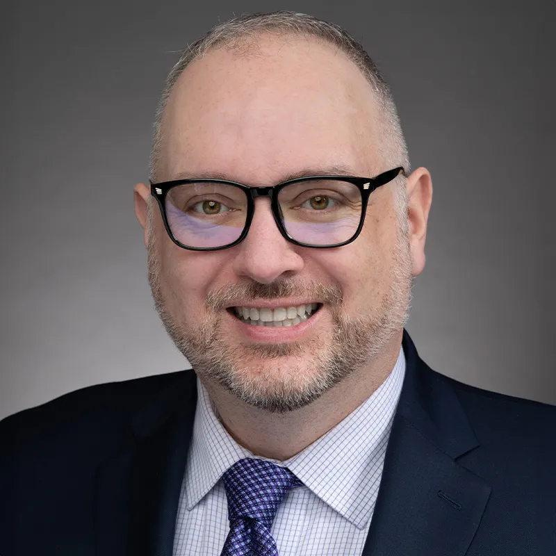 A portrait of a man with a short hair, light skin, and a neatly trimmed gray beard. He is wearing black rectangular glasses, a dark navy suit, a white checkered shirt, and a purple patterned tie. The background is a neutral gray gradient, giving the image a professional and polished appearance. His expression is friendly and confident, with a slight smile.