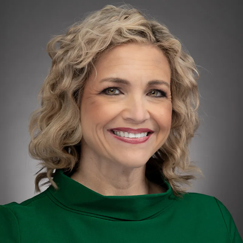 A portrait of a woman with shoulder-length, wavy blonde hair and light skin. She is smiling brightly and wearing a green top with a high neckline. The background is a neutral gray gradient, creating a professional and polished appearance. Her expression is warm and approachable.