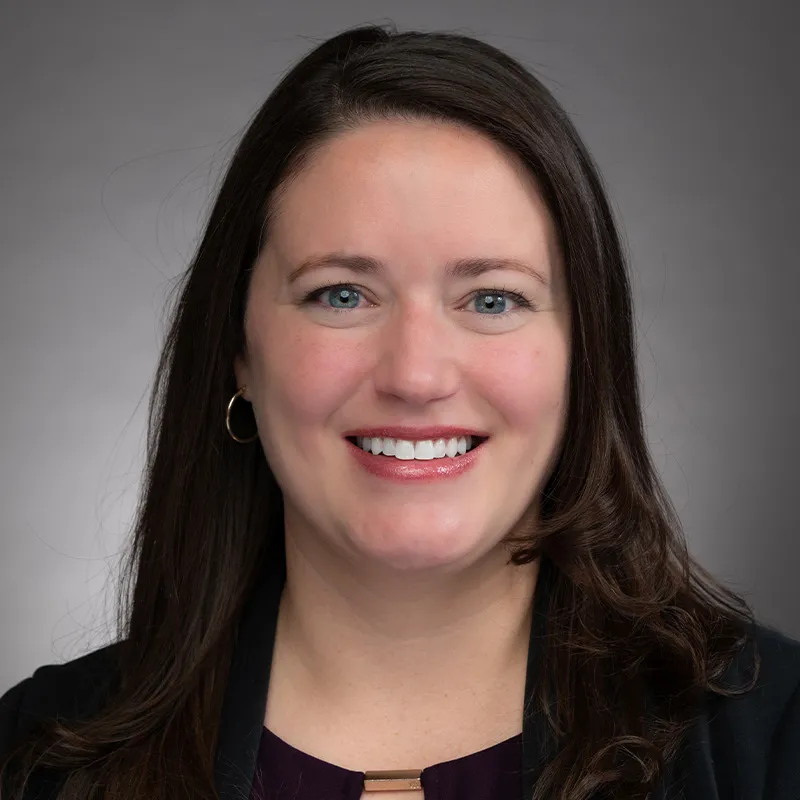 A portrait of a woman with long, straight dark brown hair and light skin. She is smiling brightly and wearing a dark purple blouse with a gold accent at the neckline, paired with a black blazer. She has small hoop earrings. The background is a neutral gray gradient, giving the image a professional and polished appearance. Her expression is warm and approachable.