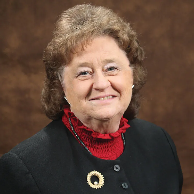 A smiling woman with short curly brown hair wearing a black blazer with a red ruffled blouse underneath, accessorized with a gold brooch. She is posed against a textured brown background.