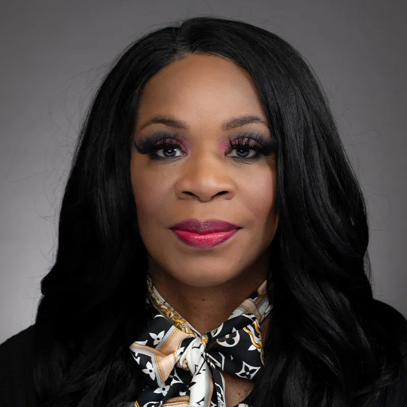 A portrait of a woman with long, wavy black hair and medium brown skin. She is wearing bold makeup with pink and purple eyeshadow, long lashes, and bright red lipstick. She has on a black blouse and a patterned scarf with gold and white details, creating a sophisticated and elegant look. The background is a neutral gray, providing a formal and polished appearance. Her expression is confident and poised.