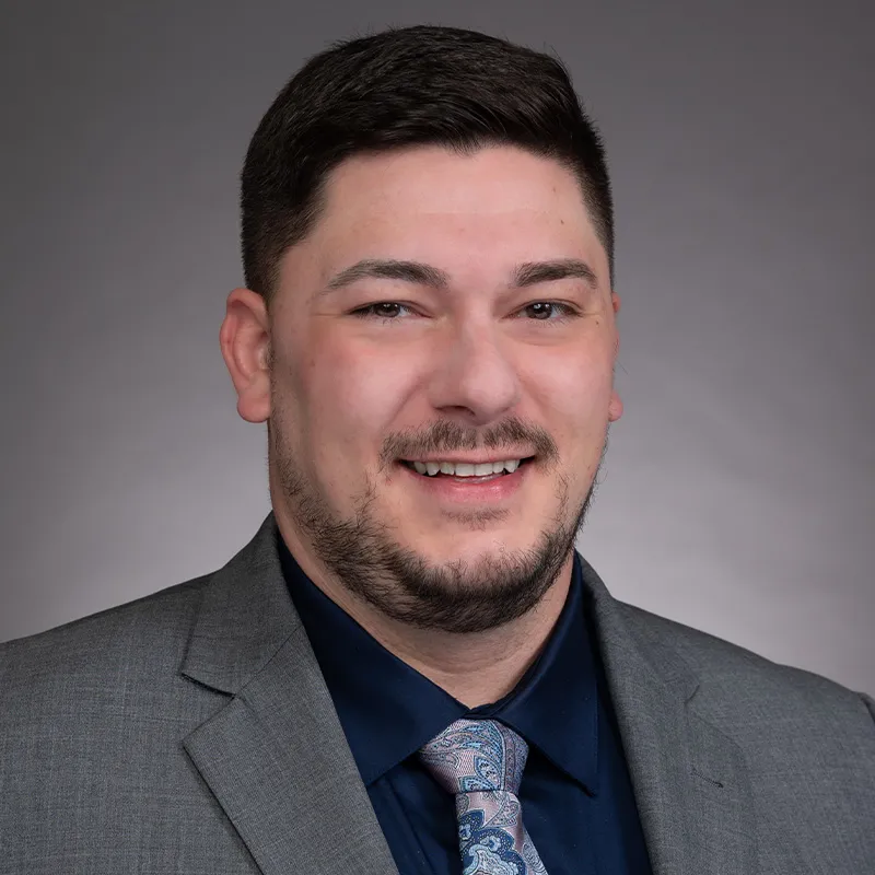 A portrait of a man with short, dark brown hair and light skin. He is smiling slightly and wearing a gray suit jacket, a navy blue dress shirt, and a paisley-patterned tie in shades of blue and pink, secured with a silver tie clip. The background is a neutral gray gradient, creating a professional and polished appearance. His expression is approachable and confident.