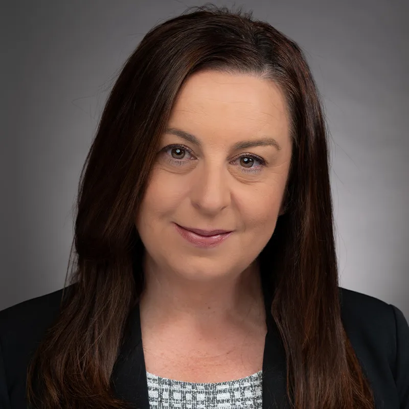 A portrait of a woman with straight, shoulder-length dark brown hair and light skin. She is wearing a black blazer over a patterned top in white and gray tones. The background is a neutral gray gradient, creating a formal and professional appearance. Her expression is composed and confident, with a slight smile.