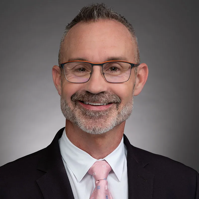 Professional headshot of a smiling man with short gray hair, a neatly trimmed beard, and glasses, wearing a black suit, white dress shirt, and a pink patterned tie, set against a neutral gray background.