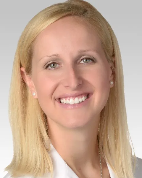 A portrait of a woman with straight, shoulder-length blonde hair and fair skin. She is smiling brightly, showing her teeth. She is wearing pearl earrings and a white top or coat, suggesting a professional setting. The background is a clean, light gray, giving the image a polished and formal appearance. Her expression is friendly and welcoming.