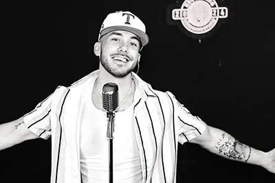 Black and white photo of a man wearing a baseball cap and striped shirt, standing in front of a microphone with arms outstretched, smiling confidently.