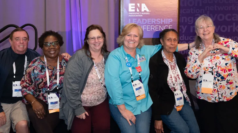 Group of smiling attendees at the ENA Leadership Conference posing in front of a purple conference banner, wearing name badges and lanyards, with one person making a heart gesture with their hands.