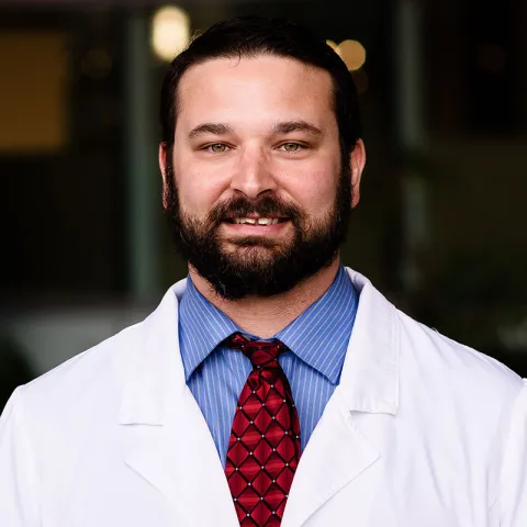 A smiling man with a full beard and short dark hair wears a white lab coat over a blue dress shirt with a red patterned tie. The background is softly blurred, suggesting an indoor professional setting.