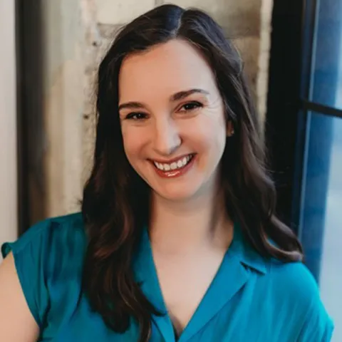 A smiling woman with long, wavy dark brown hair is wearing a teal blouse. She is positioned in front of a softly lit industrial-style window with a textured wall in the background.