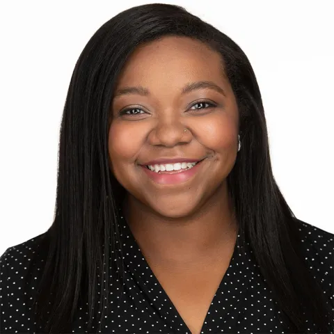 A portrait of a smiling woman with dark brown skin and long, straight black hair. She is wearing a black blouse with small white polka dots. Her nose is pierced on one side, and her makeup is subtle with soft eyeliner and natural lip color. The background is a plain white backdrop, and her expression conveys warmth and friendliness.