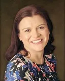 A professional portrait of a woman with shoulder-length dark brown hair. She is smiling warmly and wearing a floral-patterned top with shades of blue, red, and white.