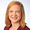 A headshot of a woman with light red hair, smiling warmly at the camera. She is wearing a professional outfit with a white background, creating a clean and polished look.