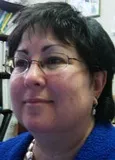 A close-up of a woman with short dark hair and glasses wearing a blue top and pearl earrings. She is in an office setting with shelves and books in the background.