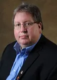 A portrait of a man with short gray hair, glasses, and light skin. He is wearing a black blazer over a blue dress shirt, with a pin visible on the blazer.