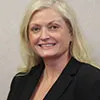 A portrait of a woman with long, straight blonde hair and light skin. She is smiling and wearing a black blazer over a light top. The background is a plain, neutral beige, creating a professional and polished appearance. Her expression is friendly and confident.