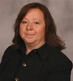 A woman with shoulder-length brown hair wearing a black blazer with decorative buttons. She has a neutral expression and is posed against a plain gray background.