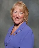 A smiling woman with short blonde hair wearing a purple blazer adorned with multiple pins. She is posed against a textured gray background.