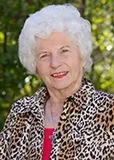 An older woman with short white hair wearing a leopard-print jacket over a red top. She is smiling and posed outdoors with a background of greenery.