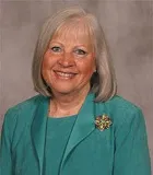 A smiling woman with shoulder-length gray hair wearing a teal blazer over a matching top, accessorized with a gold and green brooch. She is posed against a plain gray background.