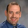 A portrait of a man with short brown hair, light skin, and a neatly trimmed beard. He is smiling and wearing a blue dress shirt with a patterned tie.