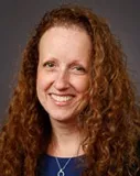 Headshot of a woman with long curly red hair, wearing a blue top and a silver necklace, smiling warmly.