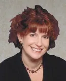 Headshot of a woman with short red hair, wearing a black top and a red beaded necklace, smiling warmly.