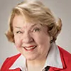 A portrait of a woman with short, styled blonde hair and light skin. She is smiling warmly and wearing a red blazer over a white collared shirt, accessorized with small earrings.