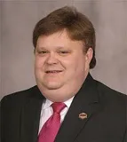 A smiling man with short brown hair wearing a black suit, white shirt, and red tie, accessorized with a small lapel pin. He is posed against a plain gray background.