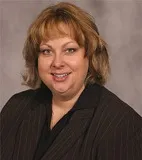 A smiling woman with shoulder-length blonde hair wearing a dark pinstripe blazer over a black top. She is posed against a plain gray background.