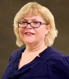 A portrait of a woman with shoulder-length blonde hair and light skin. She is wearing glasses and a navy blue blouse. 