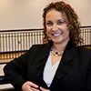 A smiling woman with curly brown hair wearing a black blazer over a white top, accessorized with a colorful statement necklace. She is posed indoors with a blurred staircase railing in the background.