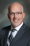 A smiling man with a shaved head and glasses wearing a gray suit, white shirt, and striped tie. He is posed against a softly blurred background with shades of gray and green.